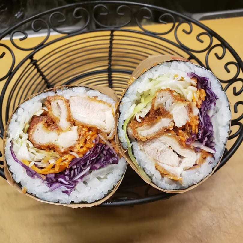 Two halves of a KANBU Sushi burrito with fried chicken, rice, and colorful vegetables in a basket.