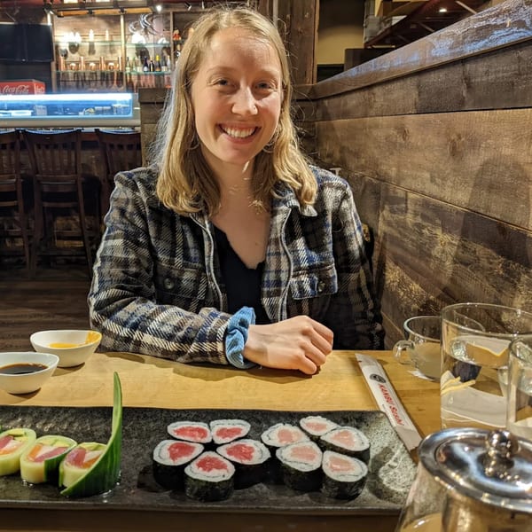 A smiling woman sits at a KANBU Sushi table with various sushi rolls and drinks.