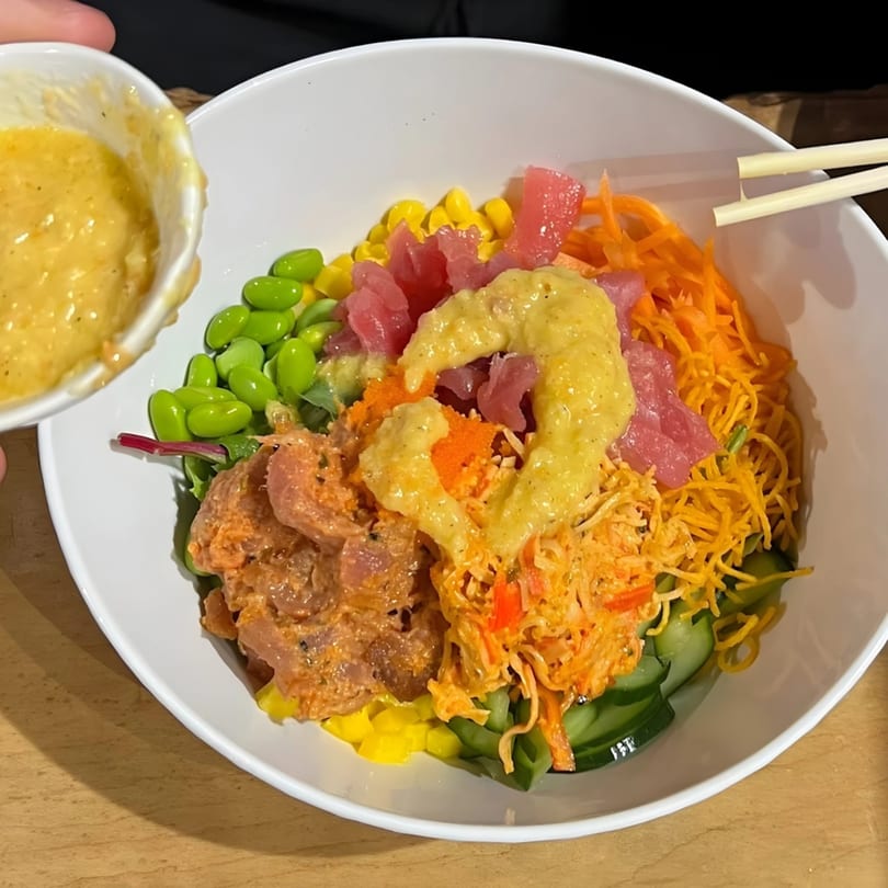 A KANBU Sushi poke bowl with tuna, edamame, corn, carrots, and sauce being poured.